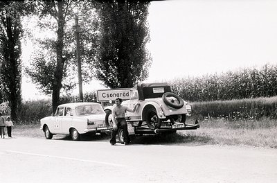 Vintage roadside scene featuring a 1960s-era white sedan towing an older classic car with a flat tire. Signpost reads "Csorar...