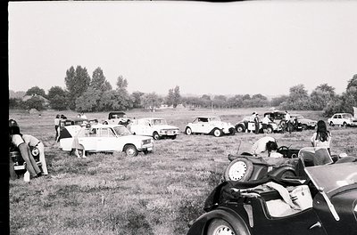 Vintage car rally in open field with mid-century compact cars and classic vehicles, likely 1960s–1970s. Participants inspect ...