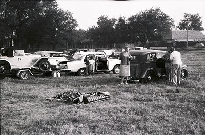 Vintage outdoor gathering featuring classic cars (1950s–1960s) parked on grass, including a trailer, station wagon, and sedan...