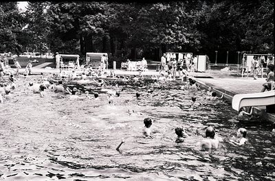 Vintage black-and-white photo of a crowded outdoor swimming pool surrounded by lounge chairs and changing cabins. Mid-20th ce...