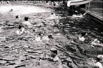 Vintage black-and-white photo of a lively seaside scene with children playing in shallow waves, mid-20th century. Wooden pier...