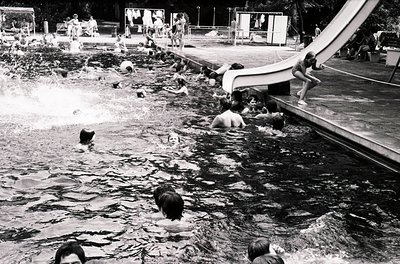 Vintage black-and-white shot of a lively outdoor pool with curved concrete edges and a diving platform. Crowded with swimmers...