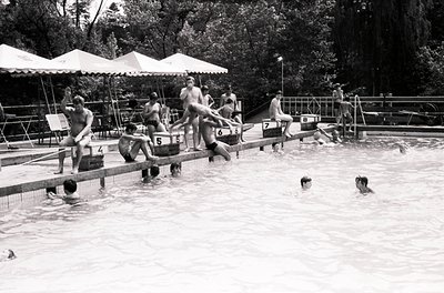 Mid-20th century outdoor swimming pool with numbered diving boards, crowded with swimmers. Wooden deck and metal railings fra...