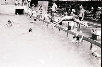 Vintage black-and-white shot of a mid-20th century outdoor swimming pool with diving board. Crowded with swimmers, children, ...