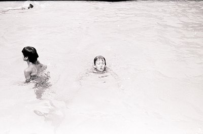 Black-and-white shot of two children swimming in shallow water, likely an indoor pool. The older child (left) appears to be t...