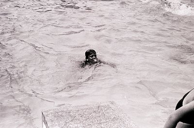 Black-and-white shot of a child swimming in shallow ocean waves, partially submerged with only head visible. Waves gently lap...