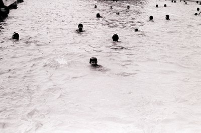 Black-and-white shot of a crowded outdoor swimming pool with numerous swimmers in shallow water, likely mid-20th century. Uni...
