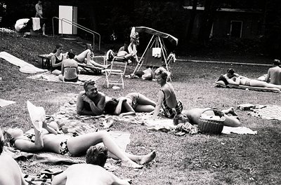 Vintage black-and-white beach scene from the 1960s–70s, featuring sunbathers in swimwear lounging on towels and inflatables. ...