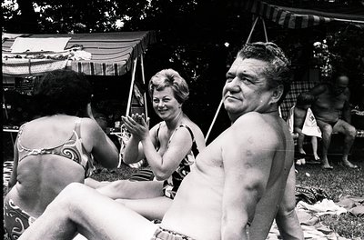 A 1960s beach scene captures three adults under a striped canopy, likely at a resort. The woman on left wears a patterned swi...