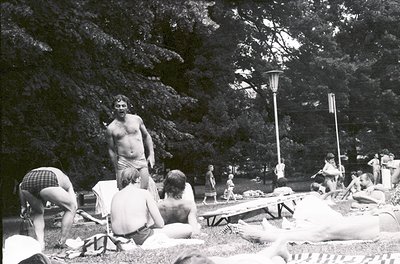 Black-and-white snapshot of a mid-20th-century outdoor gathering in a wooded park. Central figure stands shirtless, surrounde...