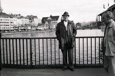 Mid-20th century man in fedora and leather jacket poses by a lakeside railing, with European-style buildings and waterfront i...