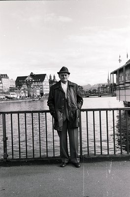 Mid-century man in a fedora and trench coat poses by a waterfront bridge, likely Zurich’s Lake Zurich. Architectural details ...