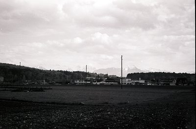 Black-and-white landscape featuring a flat, open field with sparse vegetation, likely agricultural. Distant village with low-...