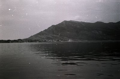 Vintage black-and-white coastal scene featuring jagged mountain range framing a calm body of water. Distinctive ripples and s...
