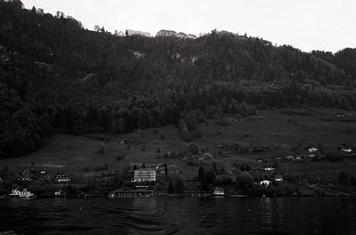 Black-and-white lakeside village nestled between forested hills and a rocky cliffside. Prominent lakeside resort with docks a...