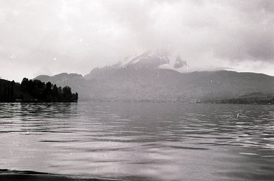 Black-and-white lake view framed by misty alpine peaks, likely European. Snow-capped mountains dominate background, with dens...