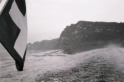 Black-and-white shot of a sailboat’s hull and sail against rugged cliffs, likely a fjord or coastal inlet. Dense forest cover...