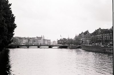 Mid-20th century urban riverscape featuring a stone bridge spanning a calm waterway. Historic European architecture with gabl...