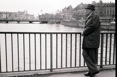 Mid-century man in a fedora and overcoat stands contemplatively on a bridge railing, overlooking a river in a European city. ...