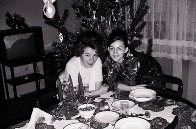 Two women pose at a festively set table in a mid-century home, likely 1950s–1960s. Decor includes a decorated Christmas tree,...