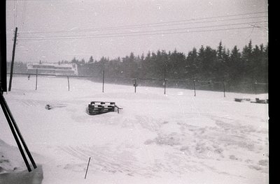 Snow-covered alpine slope with vintage sleds, likely mid-20th century. Dense forest and a multi-story building in background....