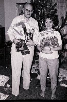 A man and woman pose indoors beside a decorated Christmas tree, holding awards and magazines. The man wears glasses, a white ...