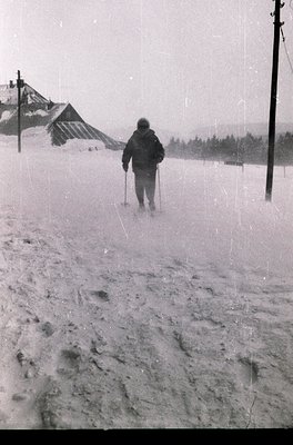 Black-and-white winter scene showing a lone cross-country skier in mid-motion on a snow-covered trail. Snow-laden trees and a...