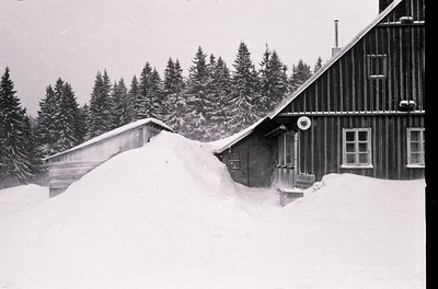 Mid-century alpine lodge buried under heavy snowdrift, showcasing classic wooden chalet architecture with sloped roof. Dense ...