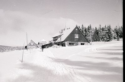 Mid-20th century alpine lodge complex blanketed in snow, surrounded by evergreen forest. Timber-framed buildings with peaked ...
