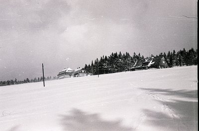 Mid-century alpine resort complex nestled in snow-covered forest, likely European. Multi-story wooden chalet-style buildings ...