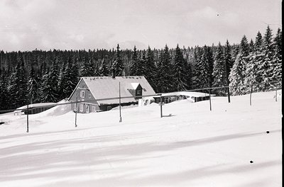 Mid-century alpine lodge nestled in snow-covered forest, likely 1950s–1960s. Two-story wooden structure with gabled roof, fla...