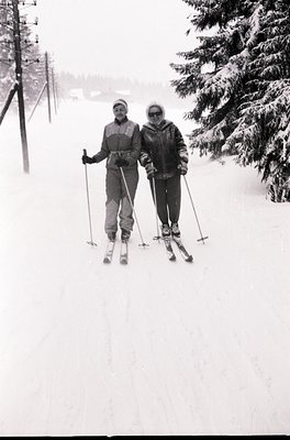 Two skiers pose on a groomed trail in mid-20th century alpine setting. Men’s and women’s ski attire from the 1950s-60s: padde...
