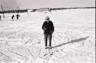 Mid-century cross-country skier pauses on groomed trail, wearing classic 1960s winter gear: knit hat, goggles, and long-sleev...