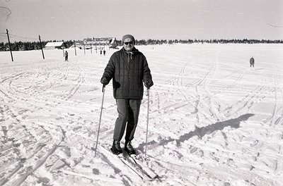 Mid-century cross-country skier in winter attire poses on groomed trail, surrounded by snow-covered landscape. Distant ski tr...