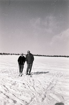 Mid-20th century cross-country skiing duo on snow-covered trail, likely or . Man in cap, woman in dark coat pose together, su...