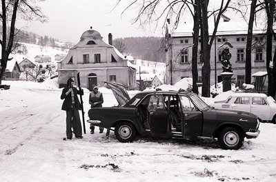 Vintage black-and-white street scene in a European town during winter, featuring a 1960s-era Zastava 750 car with open hood. ...