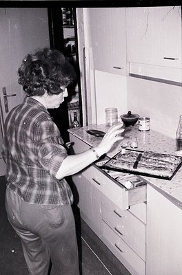 Mid-century kitchen scene featuring a person in a plaid shirt preparing food on a retro stove, 1960s-1970s. Wooden cabinets, ...