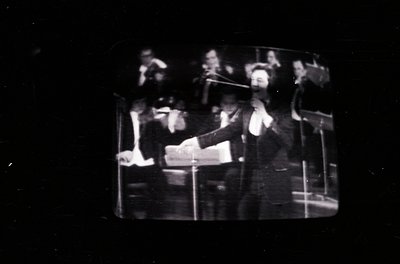 Vintage black-and-white photo of a conductor leading an orchestra, likely mid-20th century. Formal attire, sheet music, and b...