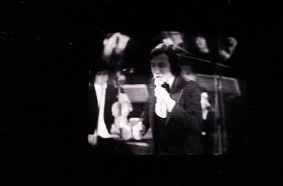 Vintage black-and-white photo of a silent film scene. A performer in a fedora and suit gestures dramatically while holding a ...
