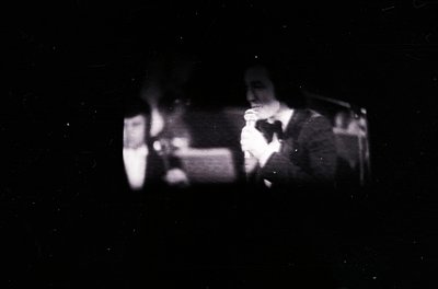 Vintage black-and-white photo of two musicians performing live, likely in a dimly lit venue. One plays guitar while the other...