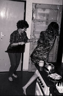 Two women in a mid-century kitchen, one adjusting the other’s patterned sweater while holding a rolled document. White stove ...