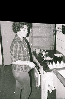 A woman in a plaid shirt and rolled-up trousers operates a vintage gas stove, cooking in a mid-century kitchen. Two mugs and ...