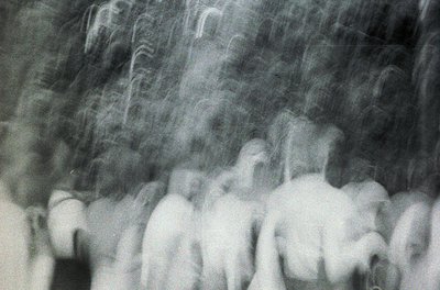 Blurred monochrome street scene with indistinct figures in motion, likely from a handheld camera. Motion blur suggests rapid ...