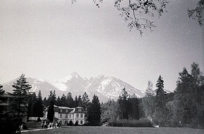 Mid-century alpine resort building nestled in a forested valley, framed by snow-capped peaks. Classic timber-framed architect...