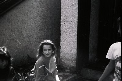 Vintage black-and-white candid of three children in an urban alleyway, likely 1960s–1970s. Girl in center wears layered cloth...