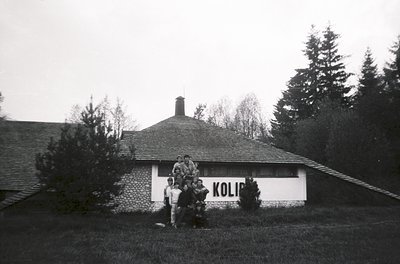 Black-and-white photo of a group posing outside a rustic stone building labeled **"KOLIB"** in the 1960s–70s. The structure f...