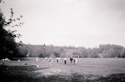 Vintage black-and-white photo of a mid-20th-century outdoor soccer game in a grassy field. Players in casual 1950s-60s attire...