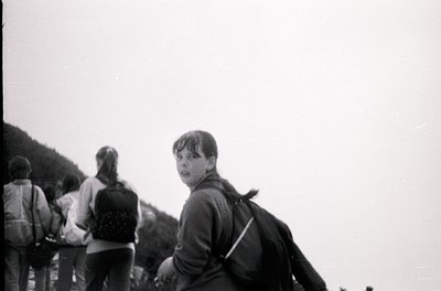 Black-and-white candid of three hikers ascending a forested trail, likely mid-20th century. Foreground subject wears a dark j...