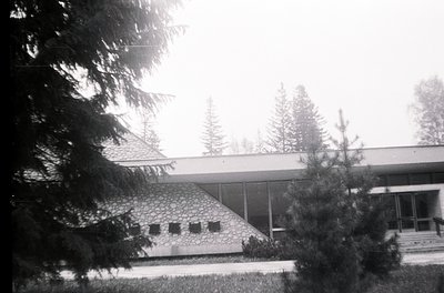 Mid-century modern building with flat roof and stone facade, framed by evergreen trees. Overcast sky suggests cloudy weather....