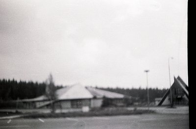 Vintage black-and-white shot of a modest, single-story building with a peaked roof in a forested area. Likely a rural or subu...